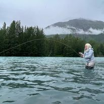 An angler stands on the shore of the Kenai River in Alaska, casting a fishing line into the turquoise water.